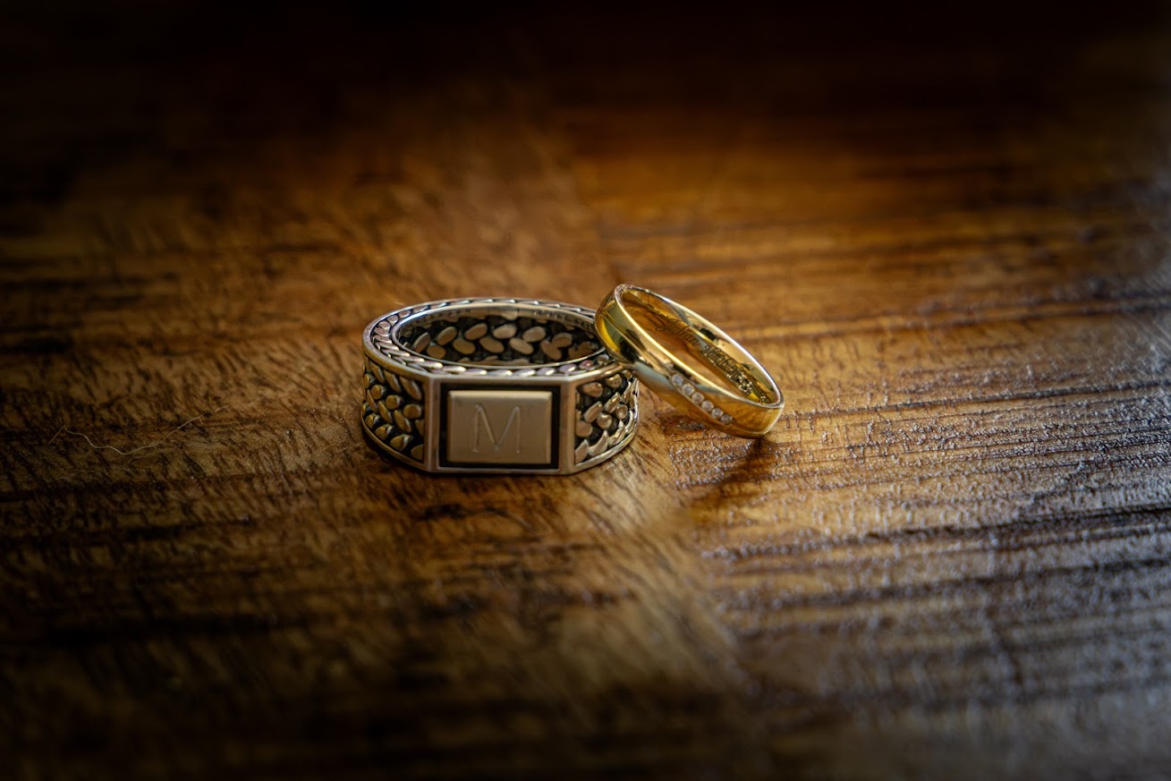Twee trouwringen op een rustieke houten ondergrond: een brede zilveren ring met de letter M en een smalle gouden ring met steentjes