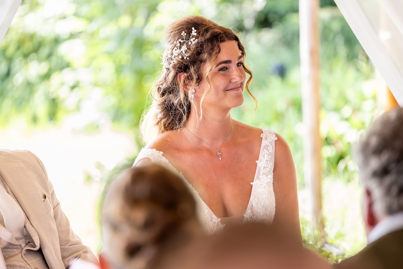 Bruid met een parel-haarsieraad in haar haar en een kanten witte jurk, glimlachend tijdens de ceremonie onder de tent