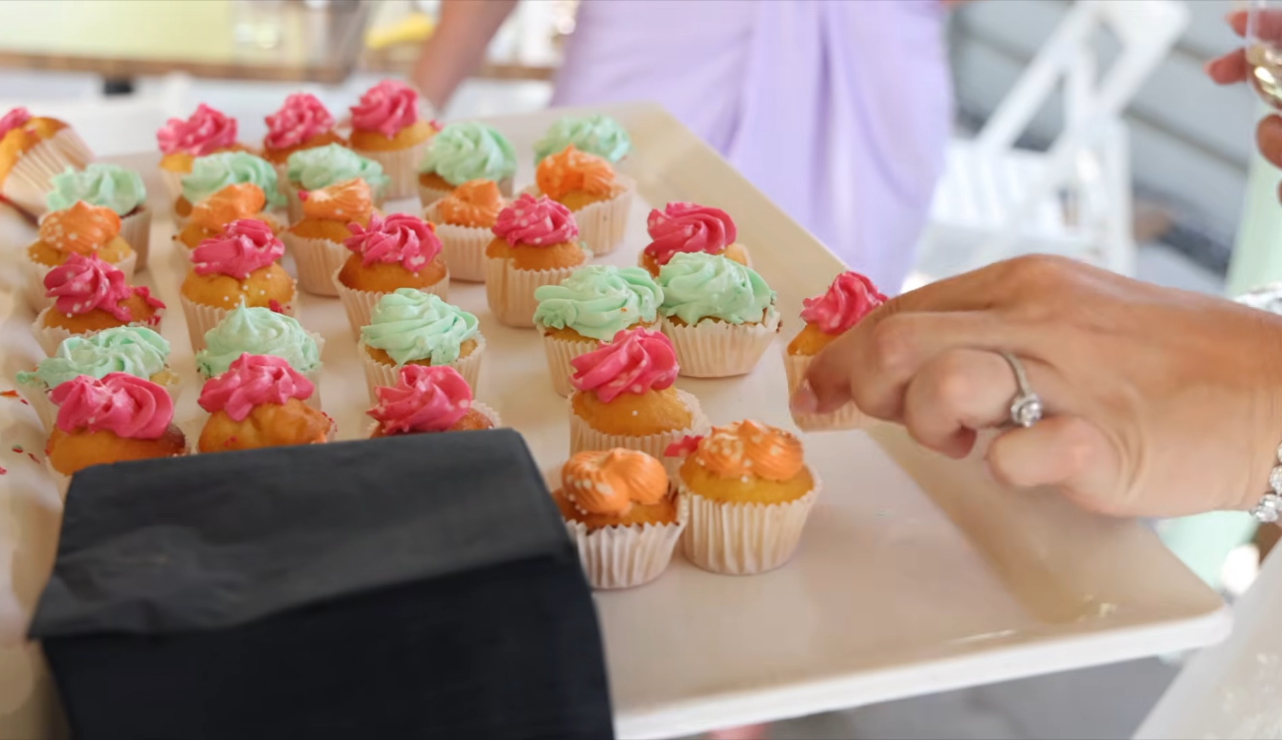 Hand met een verlovingsring pakt een mini cupcake met roze frosting van een dienblad